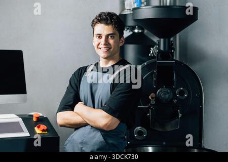 Portrait of a confident male worker posing at a production coffee machine. Smiling employee in an apron standing in a coffee shop factory. Stock Photo