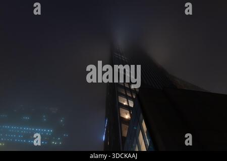 futuristic silhouette of a high-tech skyscraper standing tall against a foggy, dark blue night background. Stock Photo