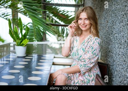Middle aged pretty woman sitting in cafeteria in dress happy smiling. Happy businesswoman girl waiting for her ordered coffee and smiling. || Model released Stock Photo
