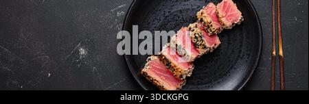 Slices of seared tuna tataki covered in sesame seeds on a dark plate, served with chopsticks on a black stone background space for text Stock Photo