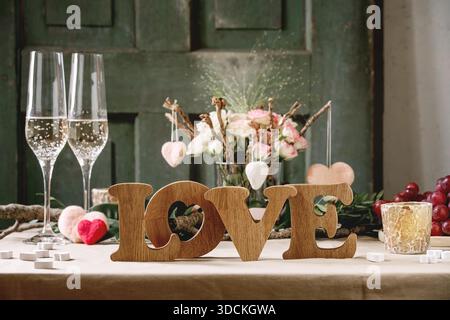 st. Valentines day or wedding romantic table setting with wooden letters love, needle felted hearts, bouquet of pink roses, couple glasses of champagne and red grapes on natural linen tablecloth. Stock Photo