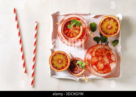 Blood orange iced cocktails in glasses, decorated by slice of oranges and fresh mint on skewers, served in ceramic tray with retro straw over white marble background. Flat lay, space. Stock Photo