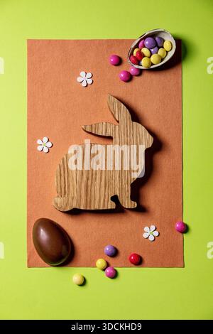 Easter greeting card with wooden decorations, chocolate sweets and eggs, daffodils on bright felt orange green background. Flat lay, copy space. Stock Photo