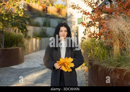 Beautiful woman with curly dark hair holding yellow maple leaves, standing by a brick wall with red ivy. Autumn sunlight highlights the warm and cozy Stock Photo