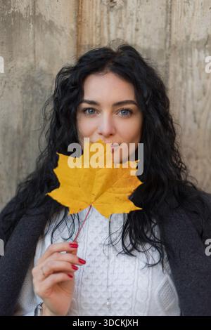 Pretty woman model with dark curly hair holding a yellow maple leaf over face, wearing a white sweater and gray coat, standing against a concrete wall Stock Photo
