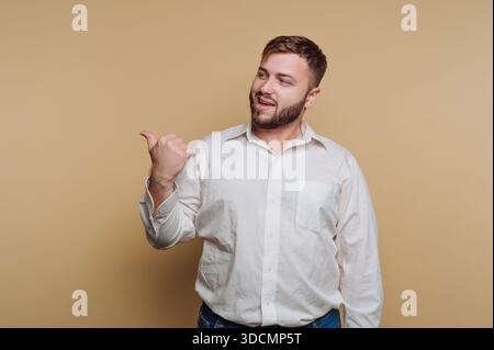 A man stands against a simple light-colored background. He smiles and points his thumb up. His casual look suggests a friendly atmosphere. Stock Photo