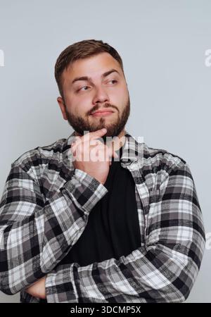 A man stands indoors with his arms crossed. He has a thoughtful expression on his face while looking slightly up. He wears a plaid shirt over a black Stock Photo