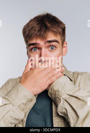 A young man looks shocked while covering his mouth with both hands. He stands in a simple indoor space with a plain background. His eyes are wide open Stock Photo