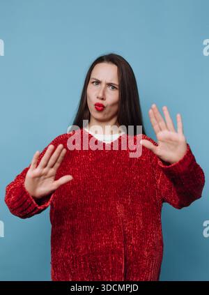 A woman stands in front of a blue backdrop, wearing a red sweater. She makes hand gestures and has a playful expression on her face. The studio settin Stock Photo