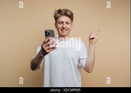 A young man stands indoors, holding a smartphone in one hand and pointing with the other. He smiles while looking at the camera. The background is pla Stock Photo