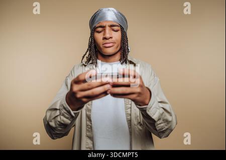 A young man stands indoors looking down at a mobile device. He has braided hair and is wearing a headscarf with a casual shirt. He appears to be conce Stock Photo