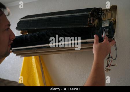 Pensive technician carefully disassembles and repairs wall-mounted air conditioning unit, using screwdriver to adjust internal components Stock Photo