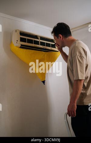 Vertical shot of focused HVAC technician using flashlight, inspecting air conditioning unit covered with yellow drop cloth during maintenance or Stock Photo