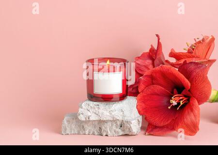 Burning red candle on stone blocks with large amaryllis flowers on soft pink background. Closeup. Stock Photo