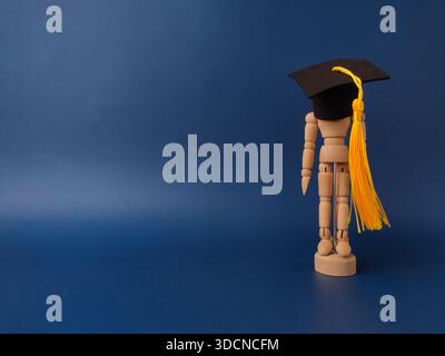 The image features a wooden artist's mannequin wearing a miniature graduation cap and yellow tassel on a blue background. Stock Photo