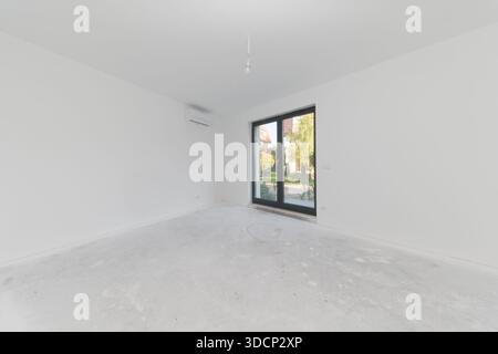Unfinished apartment interior with concrete floor and white walls. Spacious room with large balcony window and mounted air conditioning unit. New flat Stock Photo