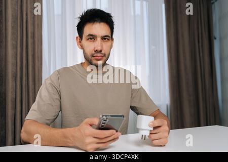 Portrait of young man holding smart socket and smartphone, looking at camera, connecting smart home device for wireless automation, setup and iot Stock Photo