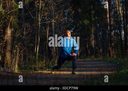 Man in athletic attire performing a deep lunge stretch on a forest trail, warming up for a workout, demonstrating fitness, healthy lifestyle, and self Stock Photo
