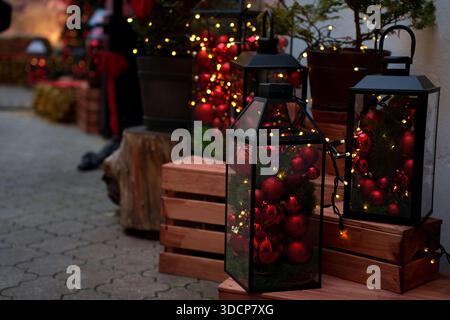 festive outdoor display featuring black lanterns filled with red christmas ornaments and string lights, situated on wooden crates, creating a warm and Stock Photo