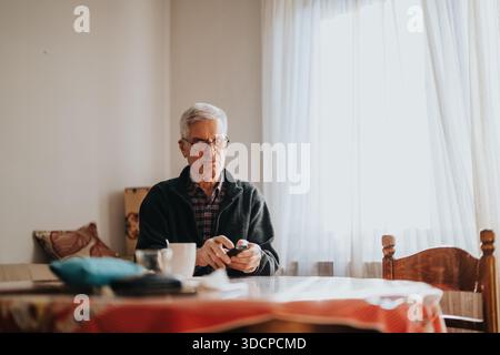 Older man sits at a sunlit dining table using a smartphone in a cozy home setting Stock Photo