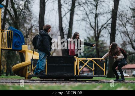 Three teenagers share laughs and conversation on a playground ride at the park Stock Photo
