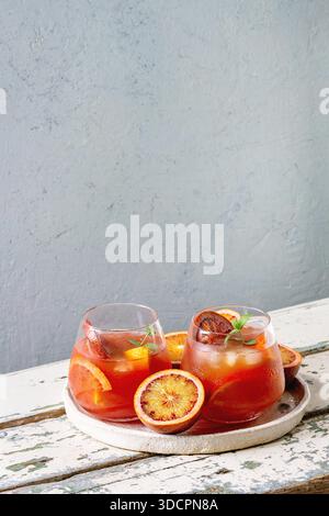 Blood orange iced cocktails in glasses, decorated by slice of oranges and fresh mint, served in ceramic tray on white wooden table with grey wall at background. Stock Photo