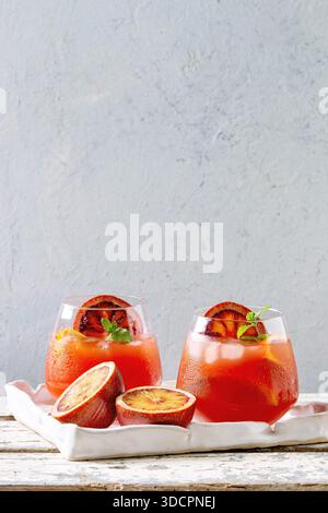 Blood orange iced cocktails in glasses, decorated by slice of oranges and fresh mint, served in ceramic tray on white wooden table with grey wall at background. Stock Photo