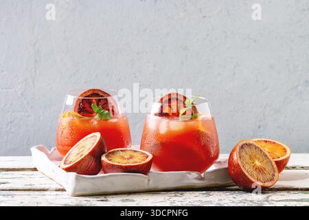 Blood orange iced cocktails in glasses, decorated by slice of oranges and fresh mint, served in ceramic tray on white wooden table with grey wall at background. Stock Photo