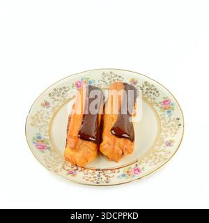 Two fresh eclairs topped with rich chocolate glaze sit on a vintage floral plate. Stock Photo