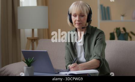 Senior old Caucasian woman in headphones writing notes watching webinar e-learning at home retired female write information looking in laptop computer Stock Photo