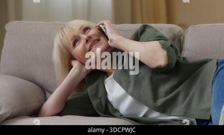 Old Caucasian woman talking mobile phone call lying on couch relaxing on sofa home cheerful smiling senior female retired lady joyful talk smartphone Stock Photo