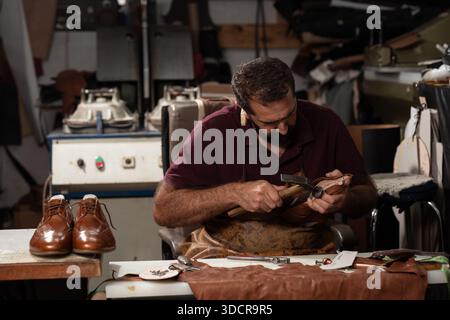 Leather shoemaker at work in a busy workshop shaping leather, with brown leather shoes on the table nearby Stock Photo