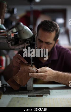 Leather craftsman sewing leather piece on an industrial machine in a busy workshop Stock Photo