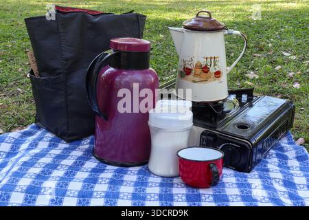 Camping gear with thermal jug enamel kettle sugar bowl and mug on portable stove. Picnic on grass with blue and white checkered tablecloth. Stock Photo
