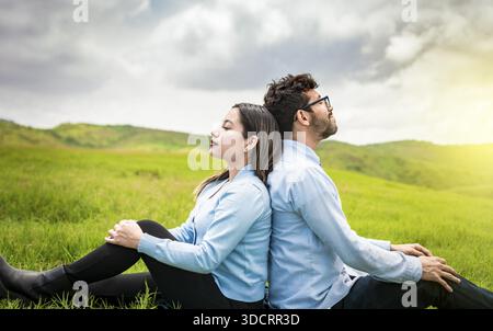 A couple sitting with their backs to each other on the grass, Wedding couple in the field sitting with their backs to each other looking towards the c Stock Photo