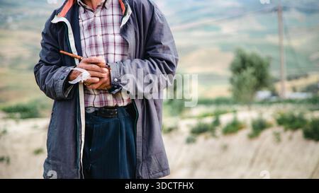 Old Moroccan man holding a traditional pipe, wearing a jacket and plaid shirt, standing in a hilly rural landscape. Stock Photo