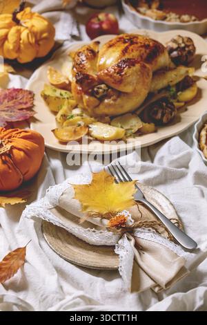 Thanksgiving or Halloween dinner with baked chicken with potatoes and lemons on big ceramic dish, pumpkin pies, plates, yellow autumn leaves and pumpkins as decorations over white table cloth. Stock Photo