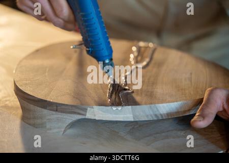 Man carpenter repairs defect on wooden stool base, craftsman using hot glue gun with liquid silicone to seal crack on wood, glue creating border for epoxy resin to fill space, material quality issues. Stock Photo