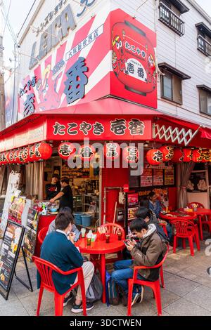 Corner Takoyaki Ichiban Osaka Sinsekaiten restaurant, Shinsekai. Large billboard with red octopus above entrance, people sitting at table with drinks. Stock Photo