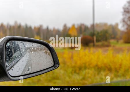 Close-up of water droplets on a car side mirror during a rainy autumn day, with a blurred rural landscape and golden trees in the background. Stock Photo