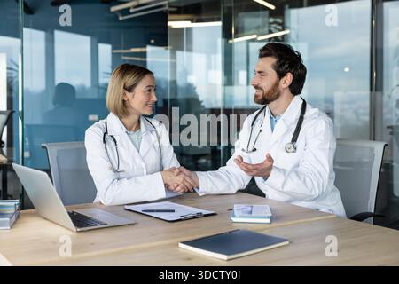 Two smiling doctors in white lab coats shaking hands, showing successful partnership and professional collaboration in a contemporary corporate medical office Stock Photo