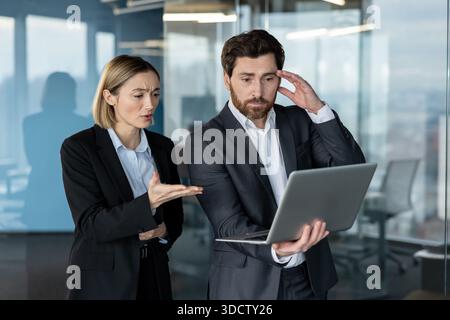 Two business colleagues in suits displaying expressions of stress and frustration while examining a laptop screen, discussing a difficult issue in a modern office environment Stock Photo