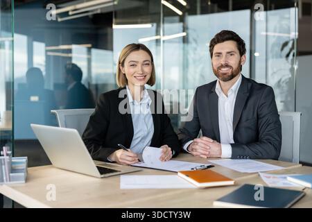 Business professionals sitting at a modern office desk, smiling at the camera while working on documents, representing collaboration, partnership, and corporate success Stock Photo