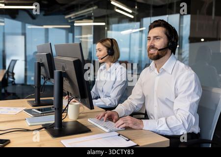 Call center agents smiling while wearing headsets, collaborating and typing on computers in a modern office, representing professional customer service and technical support Stock Photo