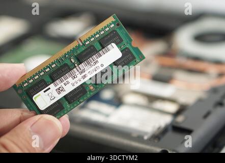 A person's hand holding a DDR3 SODIMM RAM stick during a computer repair process. Blurred laptop internals in the background indicate an ongoing hardw Stock Photo
