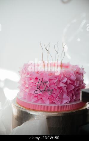 Beautiful pink ruffled birthday cake with golden candles and Happy Birthday topper against a background of balloons Stock Photo