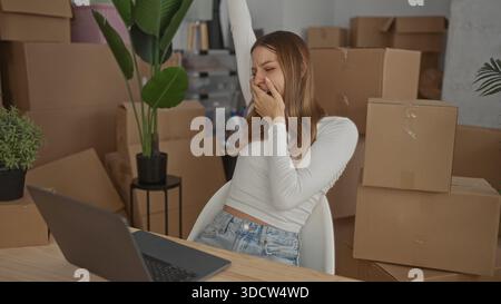 Woman stretching arms over laptop beside stacked cardboard box in a building room, leaning back in chair; moving day relief. Stock Photo
