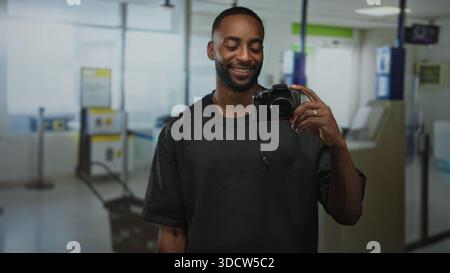 Man holds camera in mirror at airport terminal, smiling with ring visible and pendant reflected on his shirt; travel joy. Stock Photo