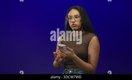 Woman holding smartphone and looking at screen, hands visible while wearing glasses and brown sleeveless shirt in a studio setting; contemplation calm Stock Photo
