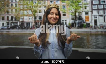 Hispanic woman wearing a gold crown smiles and beckons with both hands on amsterdam street; playful charm. Stock Photo